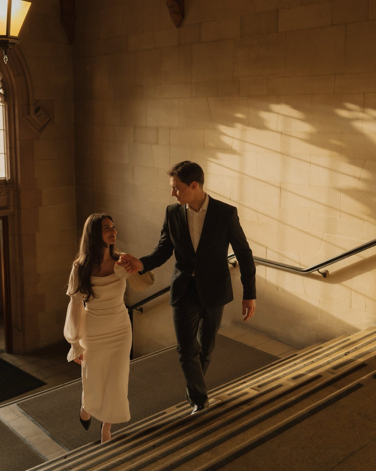 Couple in formal attire walking down stone stairs with soft, warm sunlight lighting.