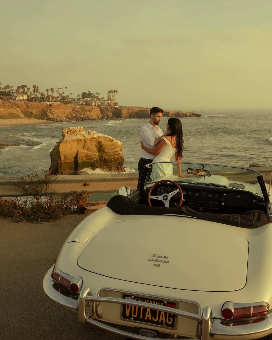 Couple embracing next to a white vintage convertible on a scenic coastal cliff