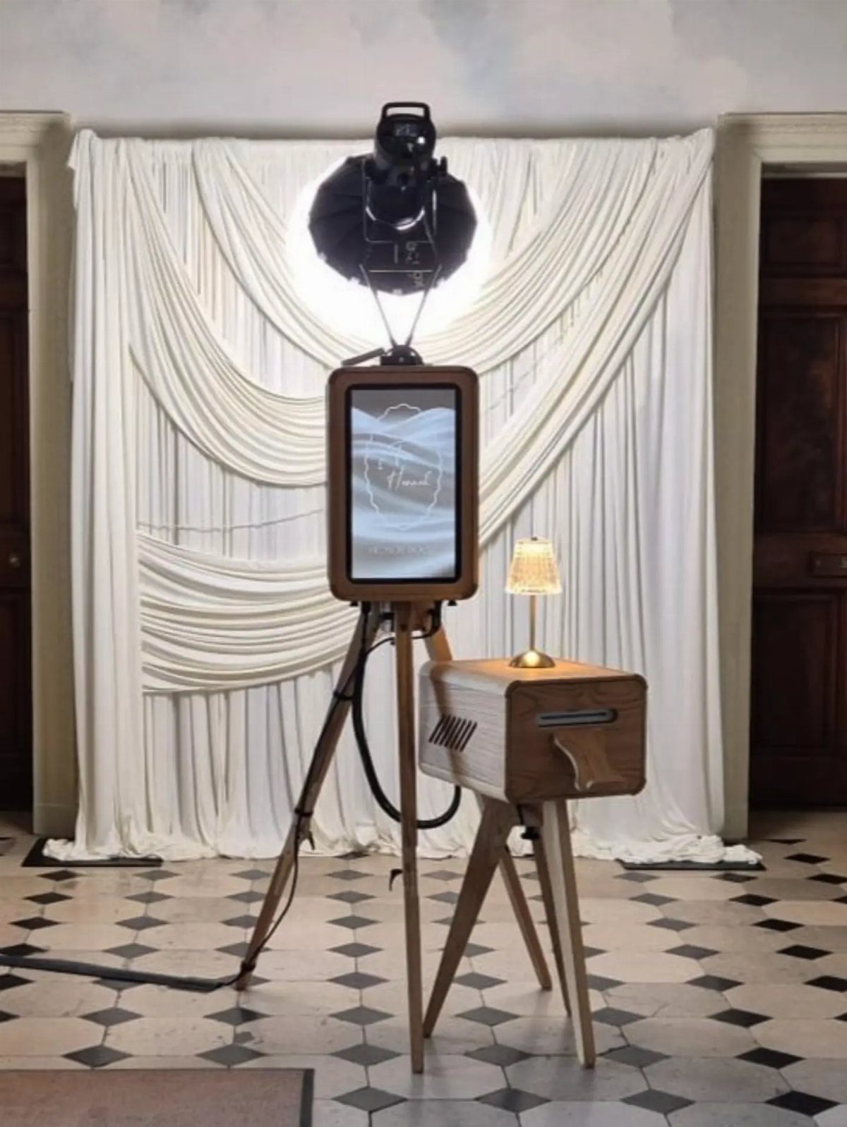 Vintage-style photo booth with wooden tripod and screen against white draped backdrop.