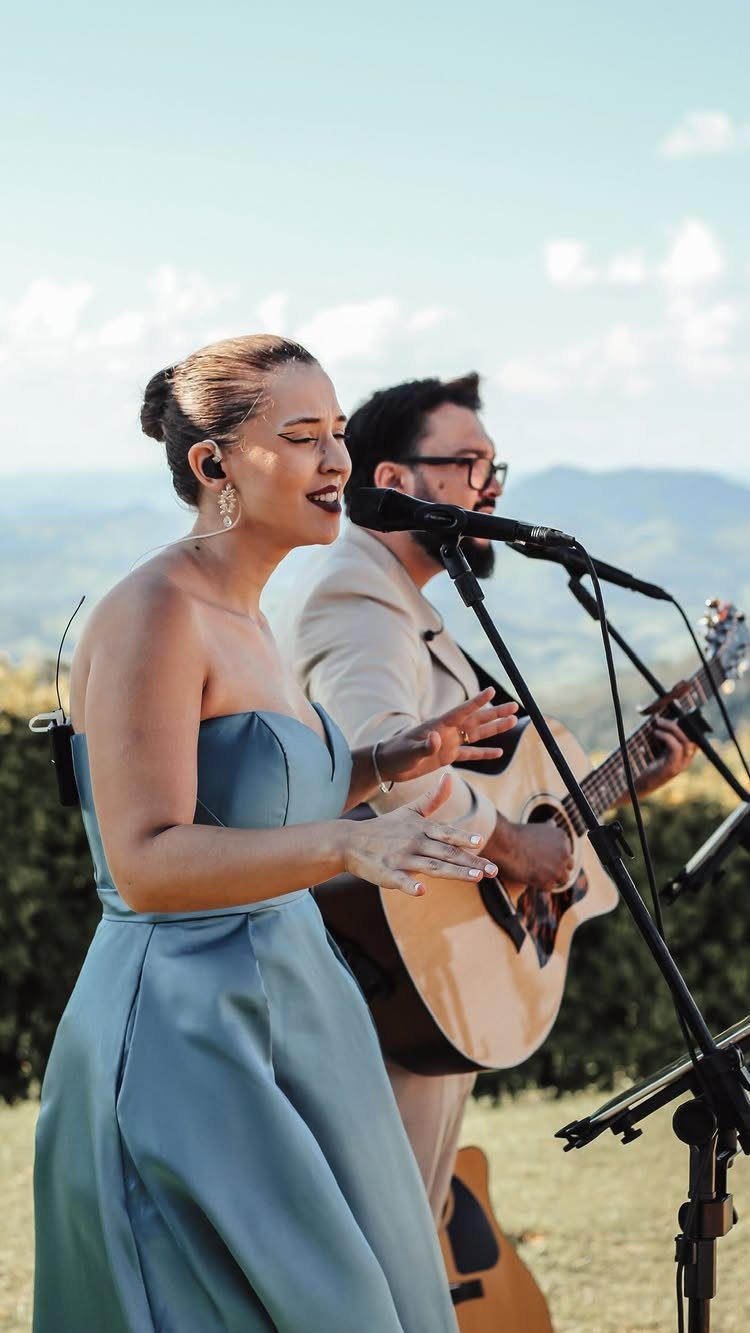 Singer in blue dress and guitarist performing outdoors with mountains in the background