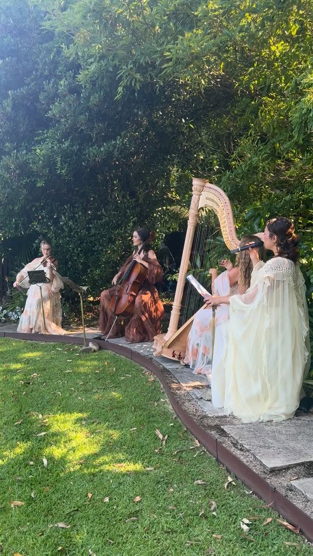 Female musicians playing harp, cello, and flute outdoors on a stone patio.