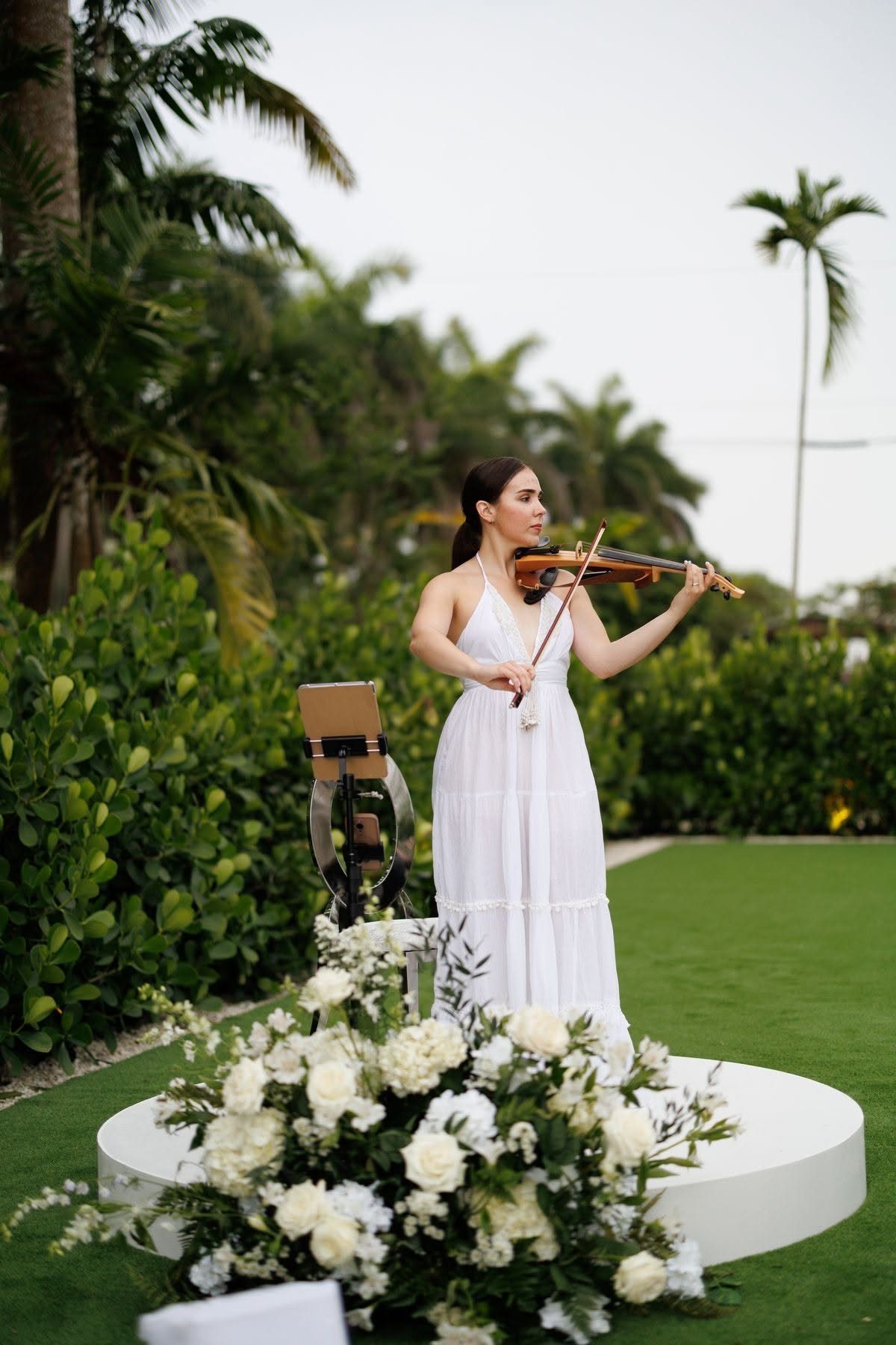 Woman in white dress playing violin behind white flowers at outdoor wedding venue