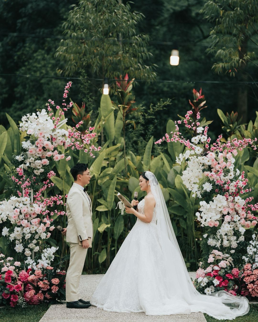Bride reading vows to groom during an outdoor wedding ceremony with floral arches.