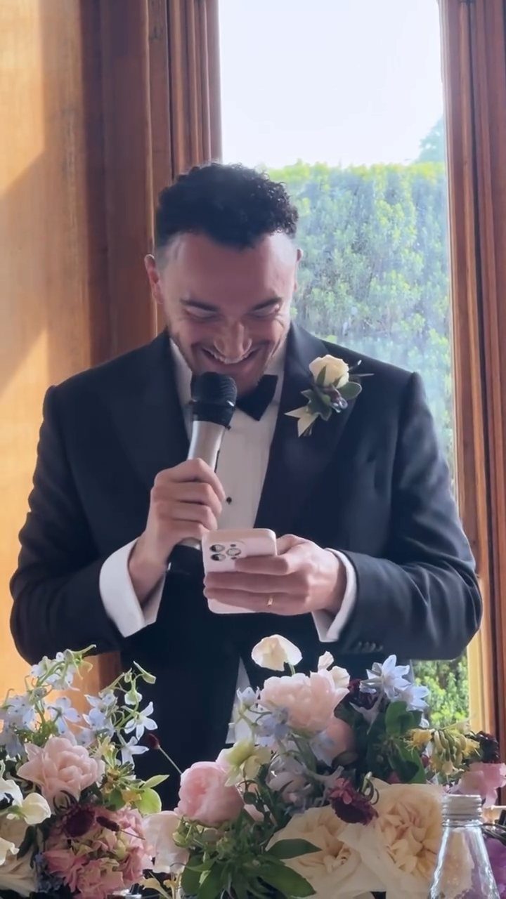 Man in tuxedo speaking into a microphone while reading from a smartphone.
