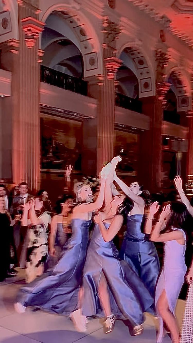 Bridesmaids in blue gowns cheering and celebrating in a grand, red-lit hall.