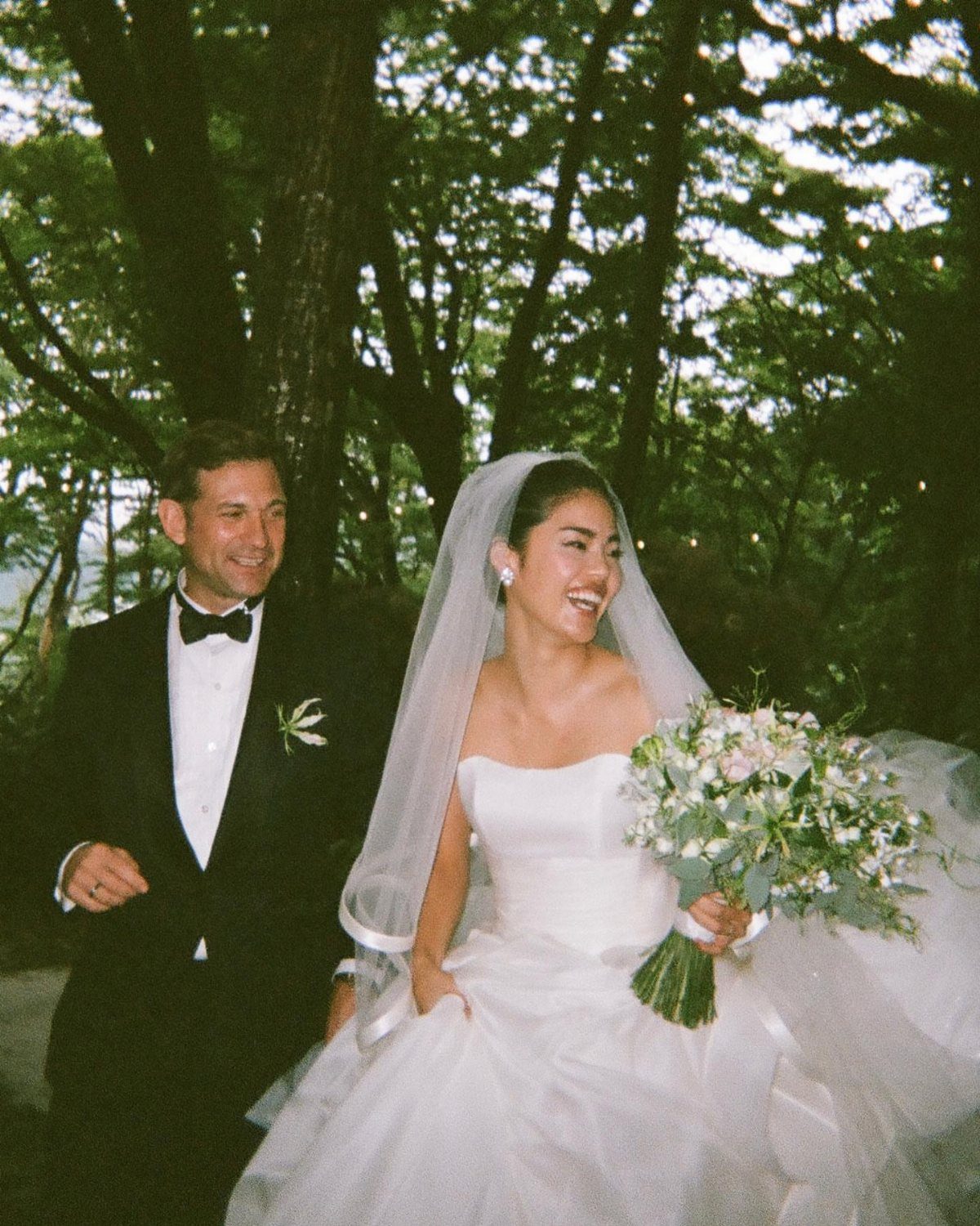Laughing bride and groom walking together through a lush green wooded area.