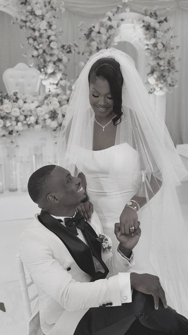 Bride in white gown and veil looking down at seated groom in tuxedo.