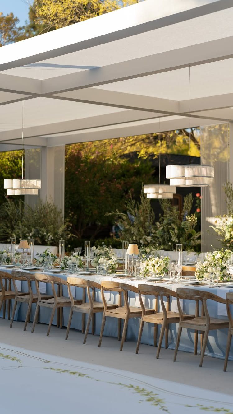 Long wedding reception table with blue linens, white flowers, and wooden chairs outdoors.