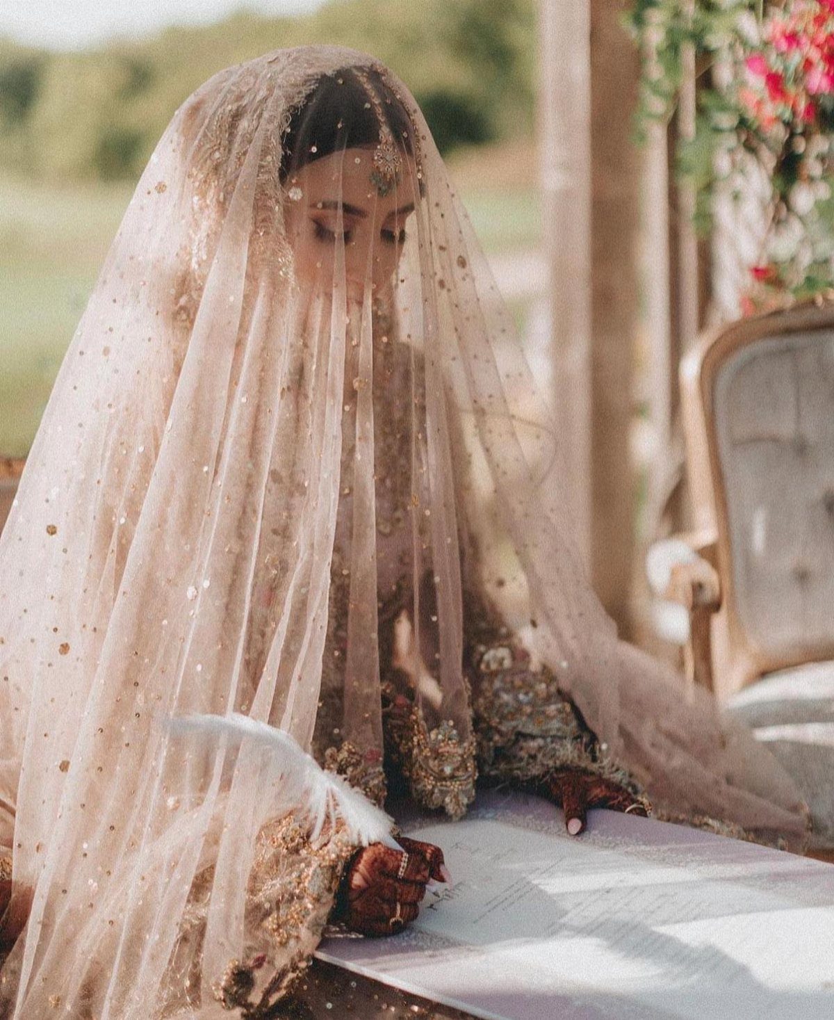 Bride in gold dress and shimmering veil writing with a white quill pen.