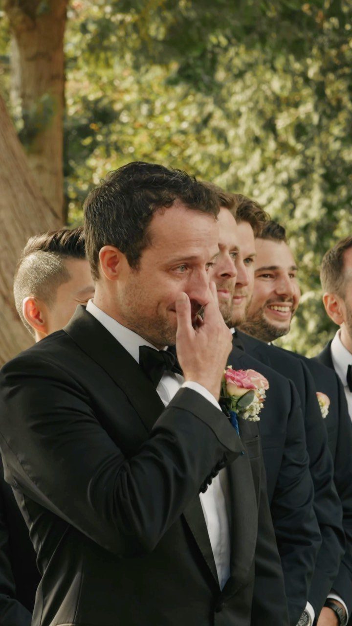 Emotional groom in a black tuxedo wiping a tear during a wedding ceremony.