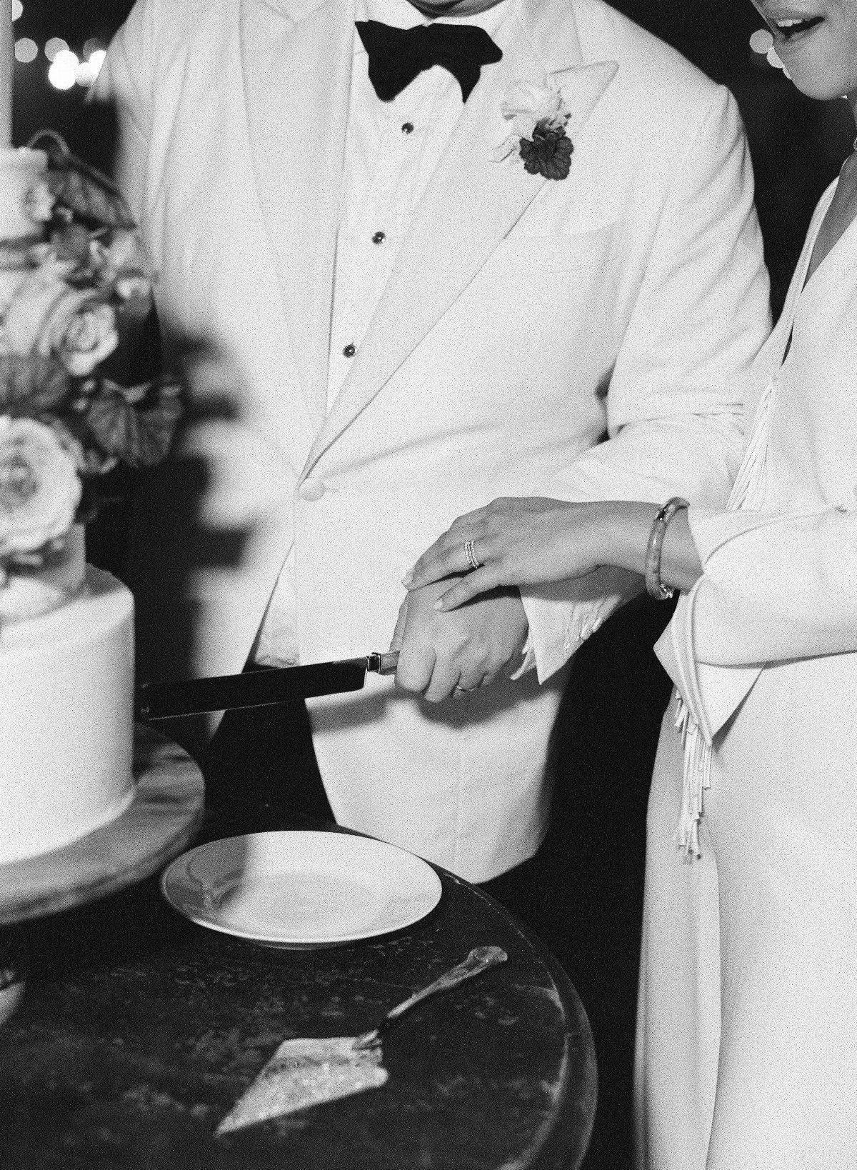 Couple in formal wedding attire cutting a tiered white cake together.