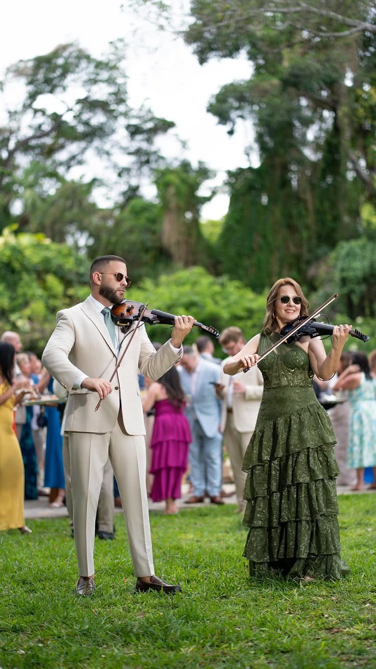 Two musicians playing violins on a green lawn during an outdoor wedding event.