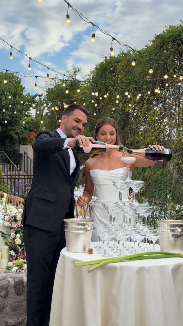 Bride and groom pouring champagne into a tower of glasses at outdoor reception.