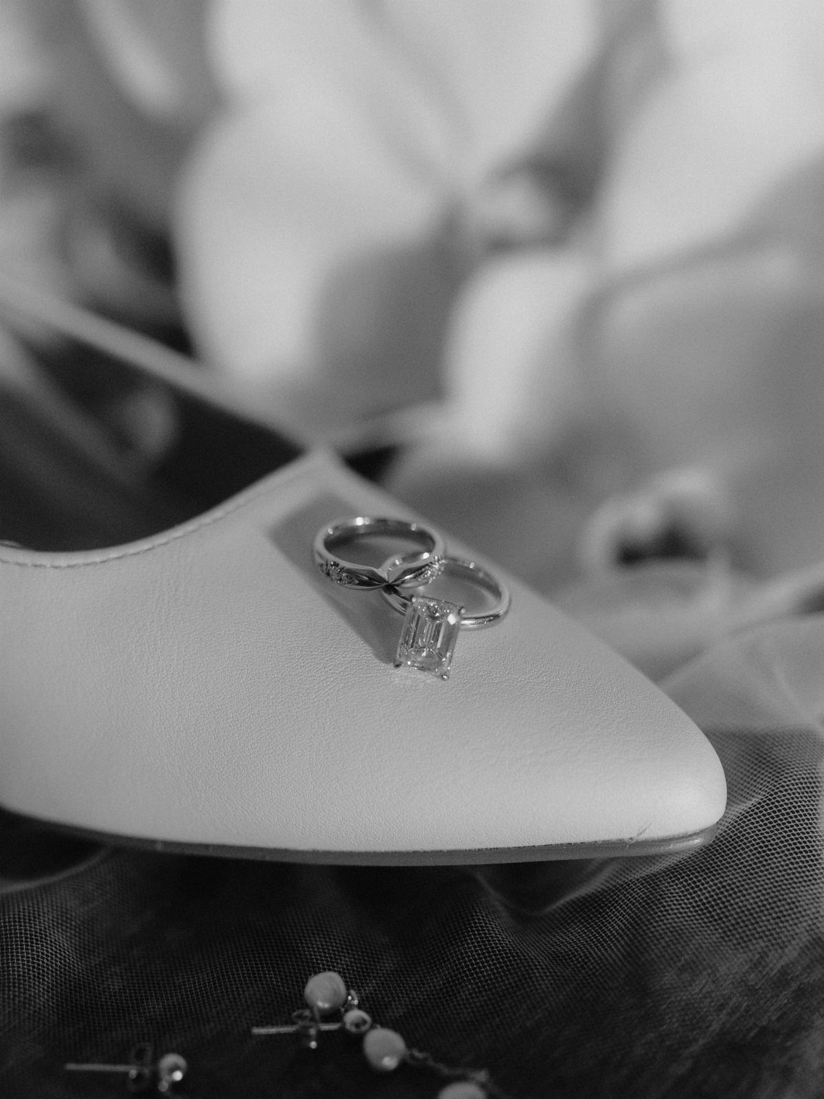 Two wedding rings resting on a white pointed-toe shoe with blurred flowers behind.