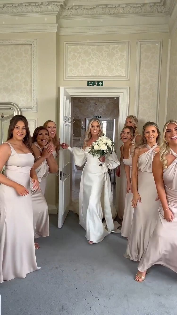 Bride in white gown standing in doorway surrounded by cheering bridesmaids in champagne dresses