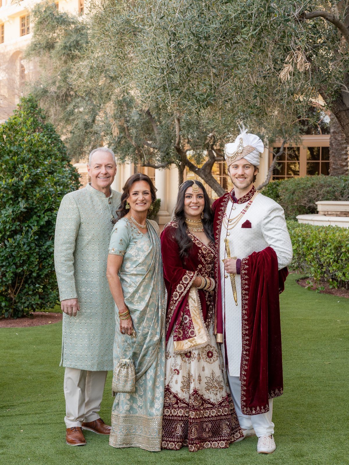 Four people in traditional Indian wedding attire posing together on a green lawn.