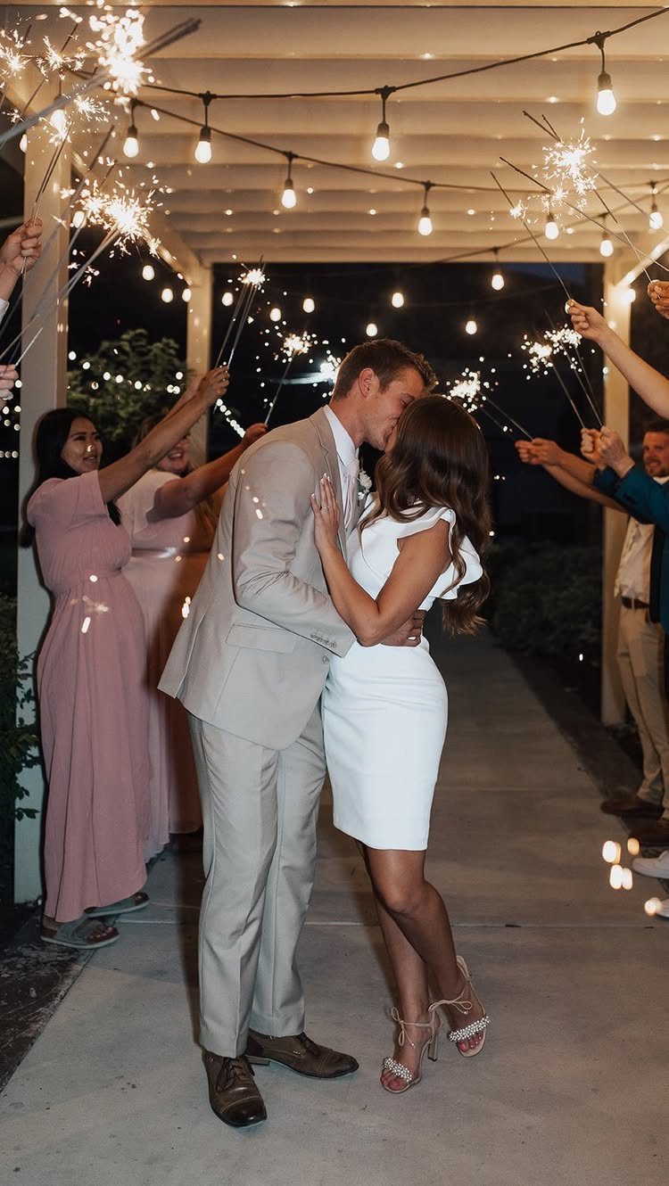 Couple kissing during a wedding send-off with guests holding sparklers under string lights.