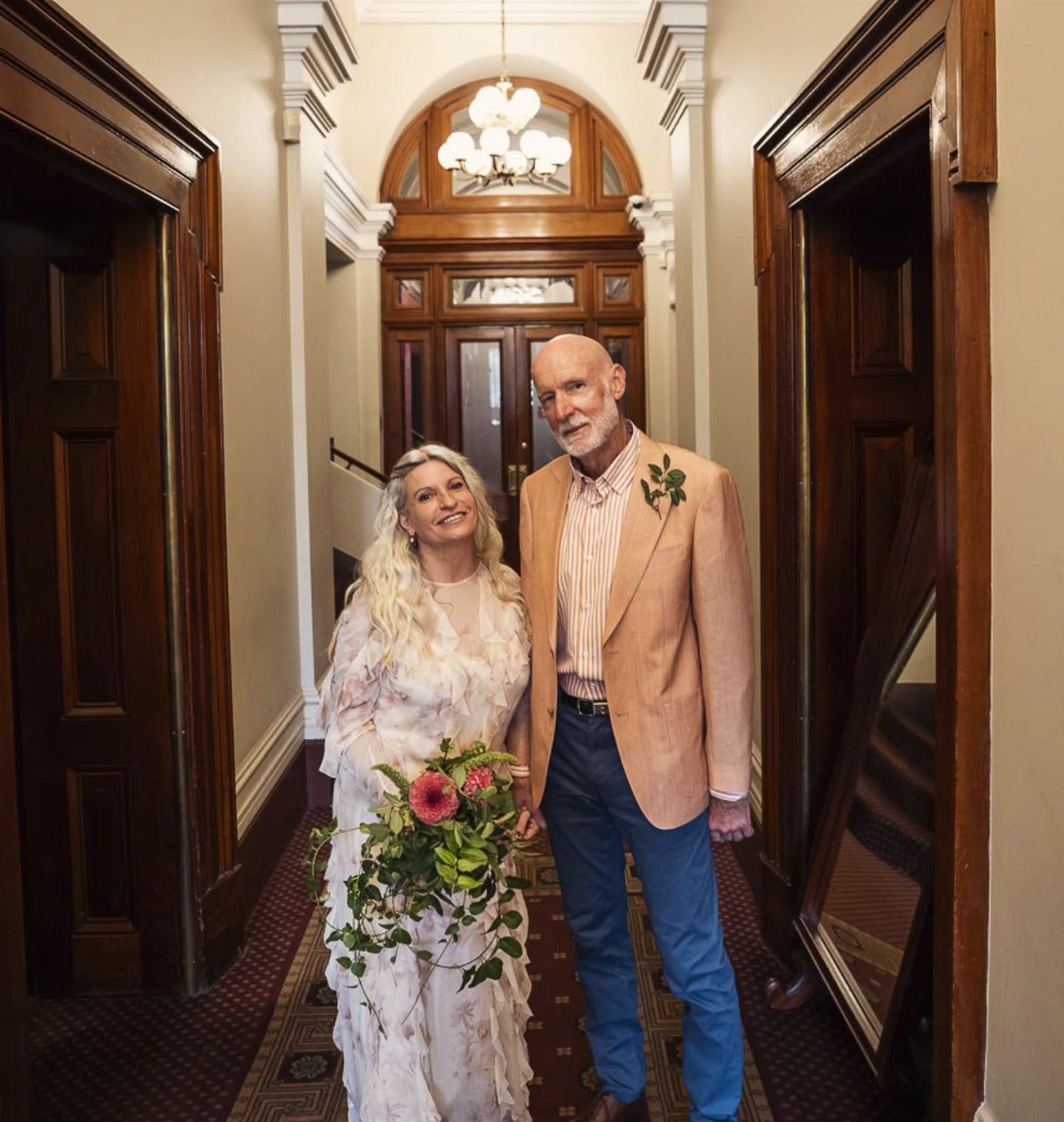 Bride in floral gown and older man in tan blazer posing in hallway