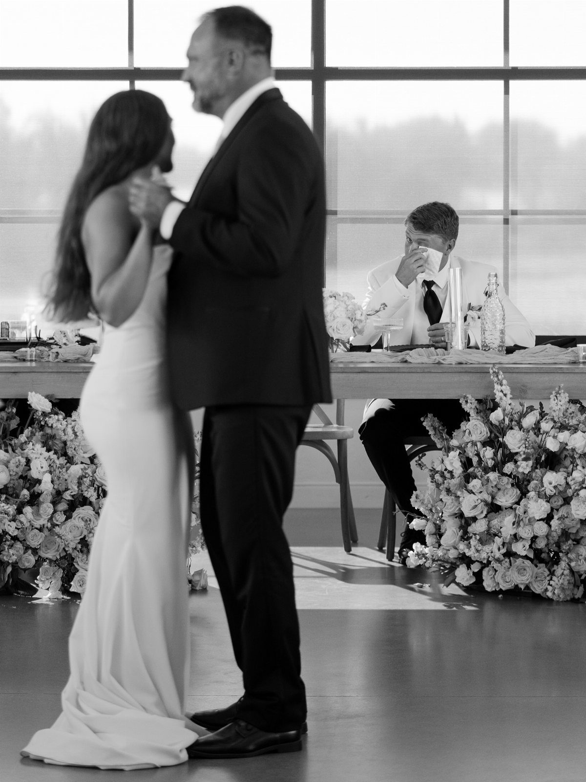Bride dancing with her father while a man watches emotionally from a table.