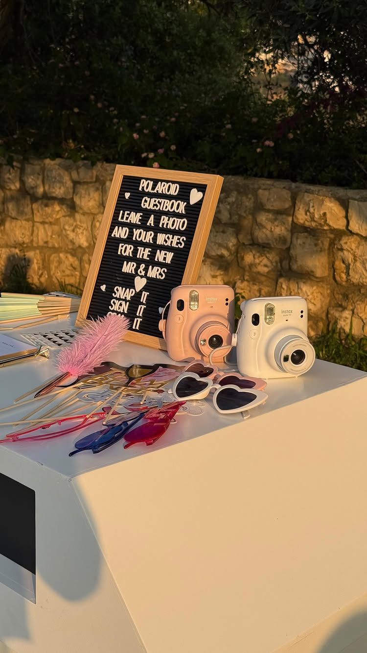 Polaroid guestbook station with pink and white cameras, heart sunglasses, and a sign.