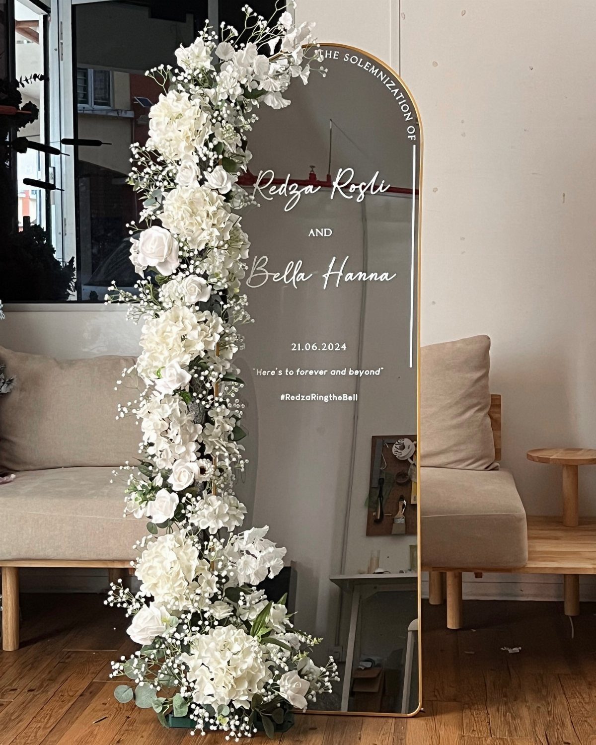 Oval mirrored backdrop with white floral arrangements and wedding text on a gold frame