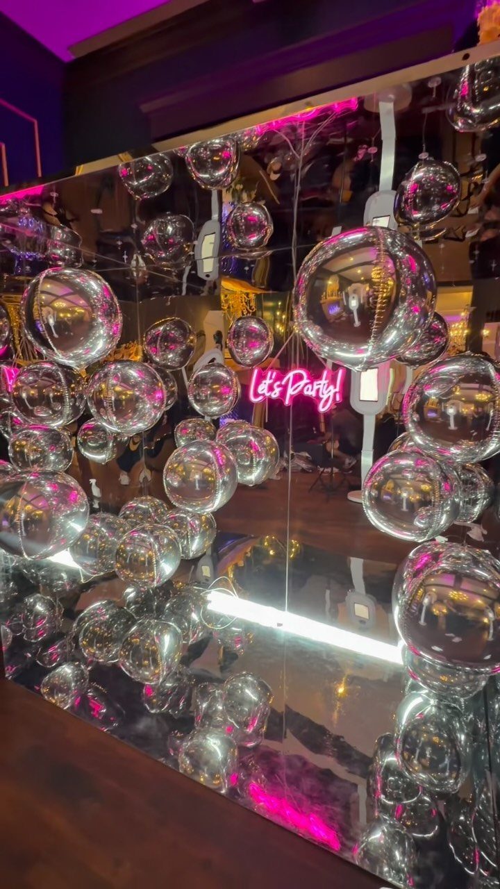 Pink Let's Party neon sign surrounded by clear silver balloons against mirrored wall