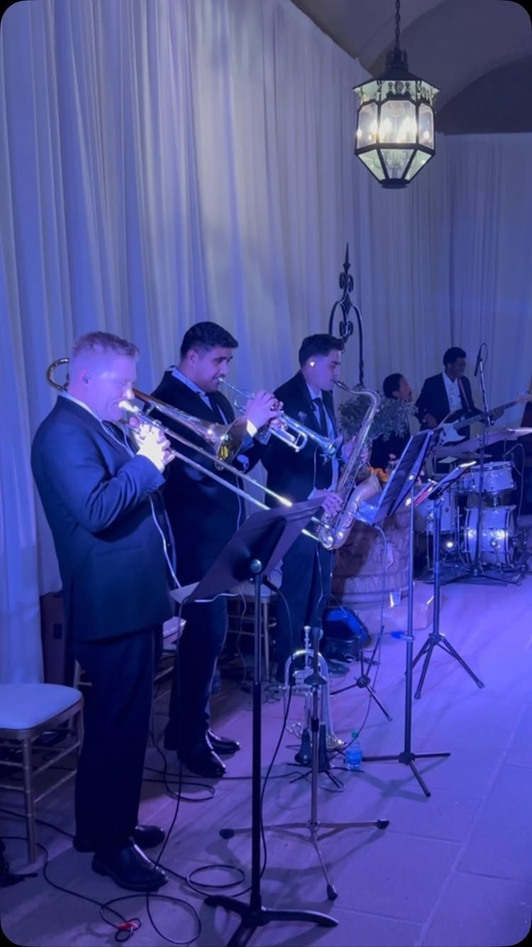 Live wedding band performing with brass instruments in a blue-lit venue with white curtains.