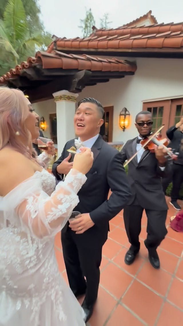 Bride and groom smiling while a violinist performs at an outdoor wedding reception
