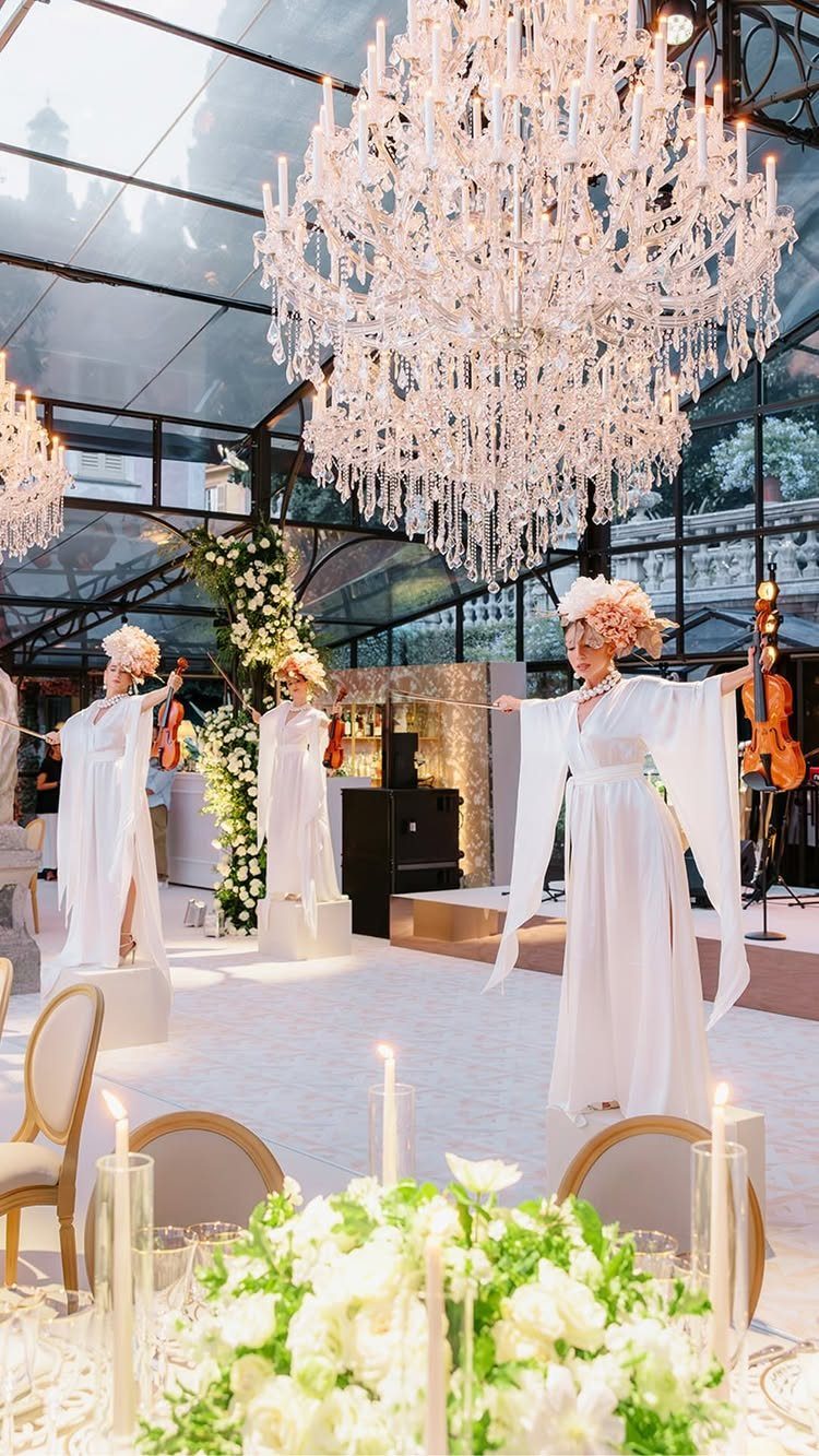 Three women in white gowns playing string instruments under a large crystal chandelier.