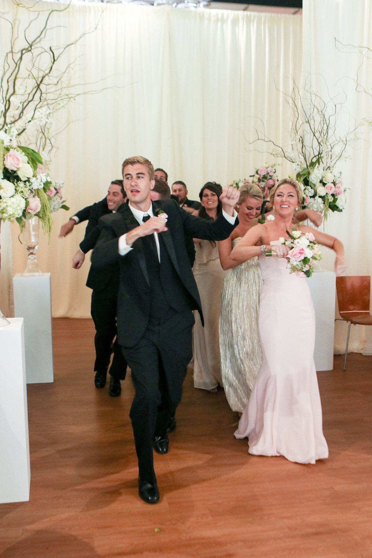 Bridal party dancing energetically into a wedding reception with white curtain backdrop