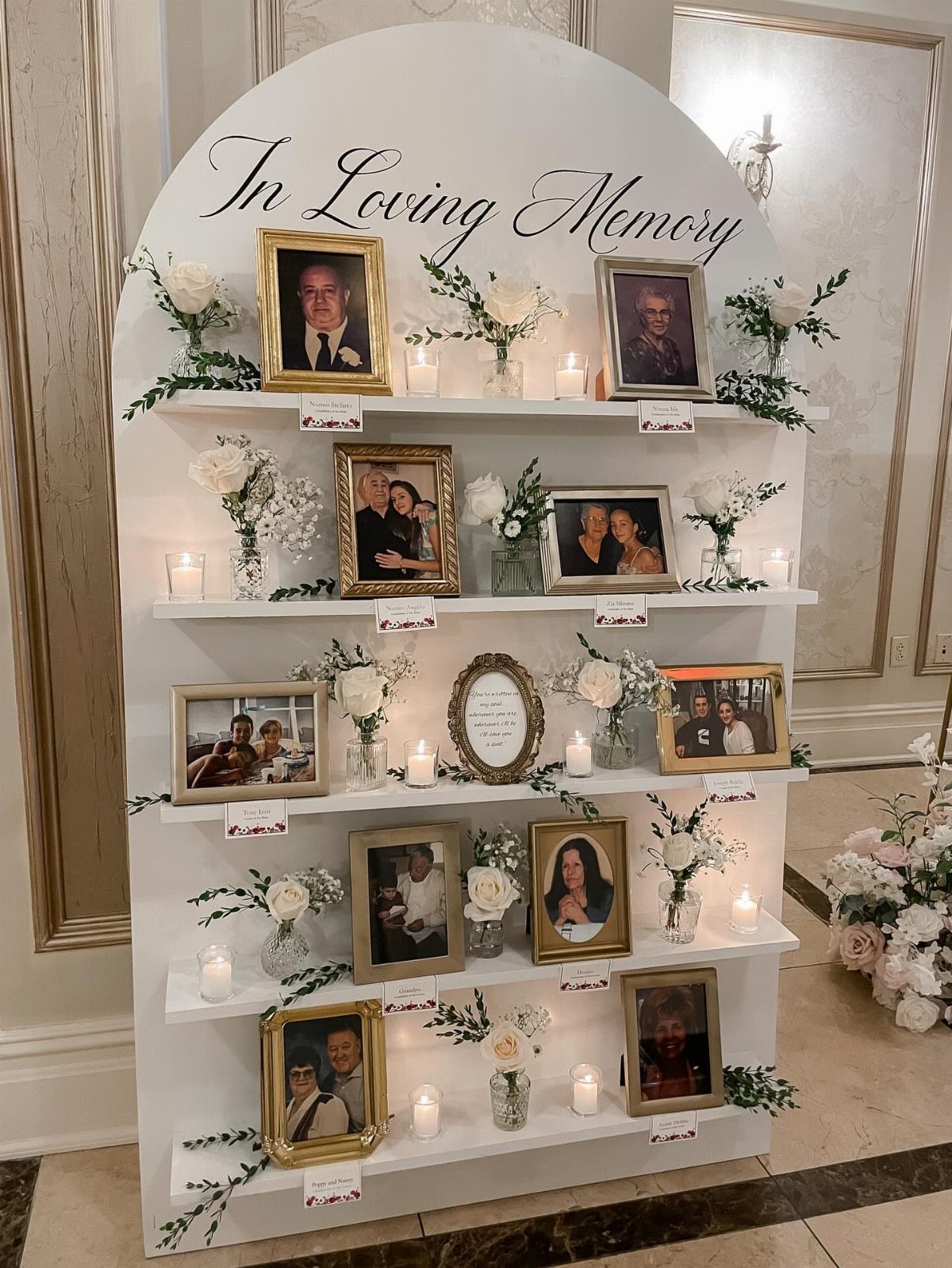 White arched memorial display with framed photos, white flowers, and candles for wedding guests