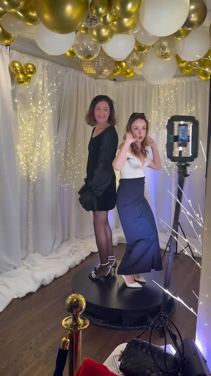 Two women posing on a 360-degree photo booth platform with gold balloons.