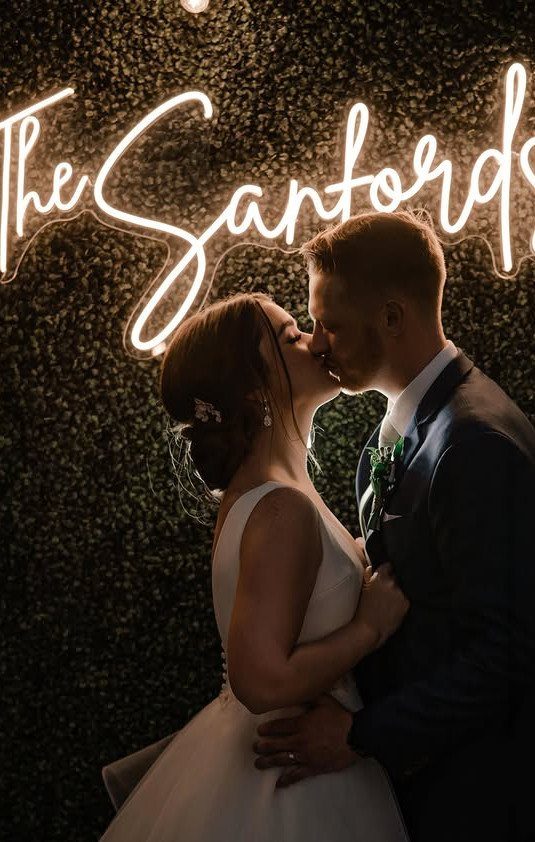 Bride and groom kissing in front of a neon sign on a hedge wall