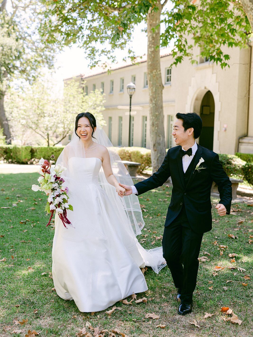 Bride and groom walking hand-in-hand across a sunny lawn in wedding attire.