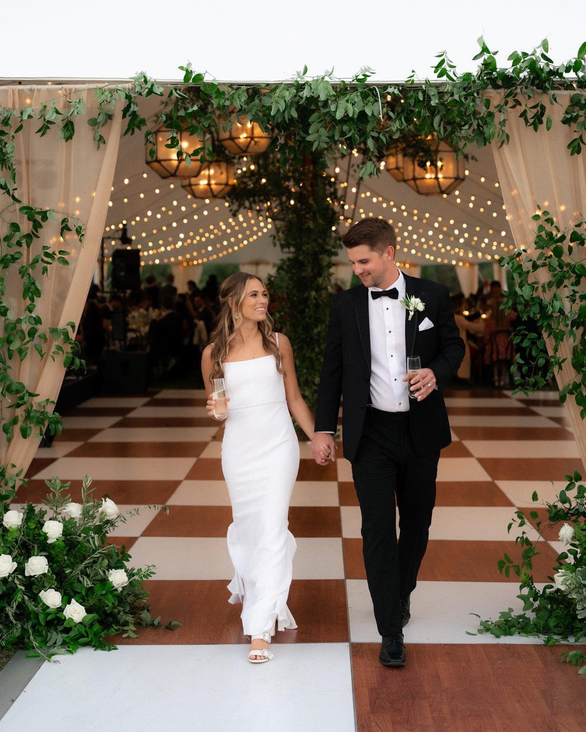 Bride and groom walking hand-in-hand across a checkered floor in a decorated tent.
