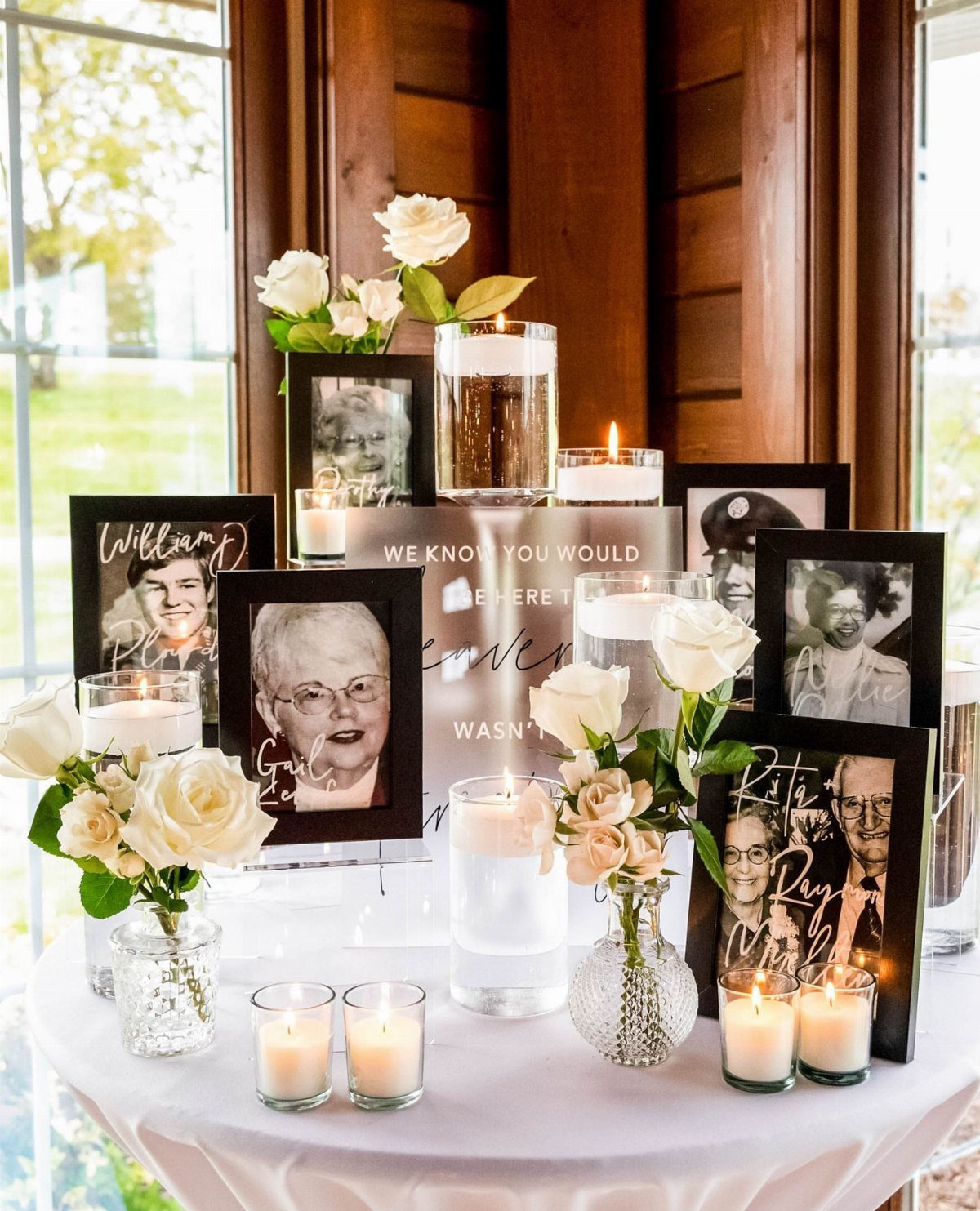 Memorial table with framed portraits, white roses, and glowing candles on white cloth