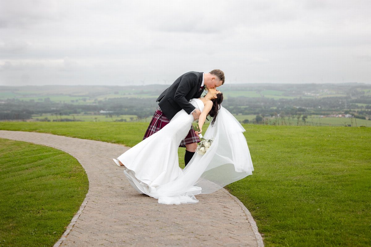 Groom in a kilt dipping bride in white gown for a romantic kiss.