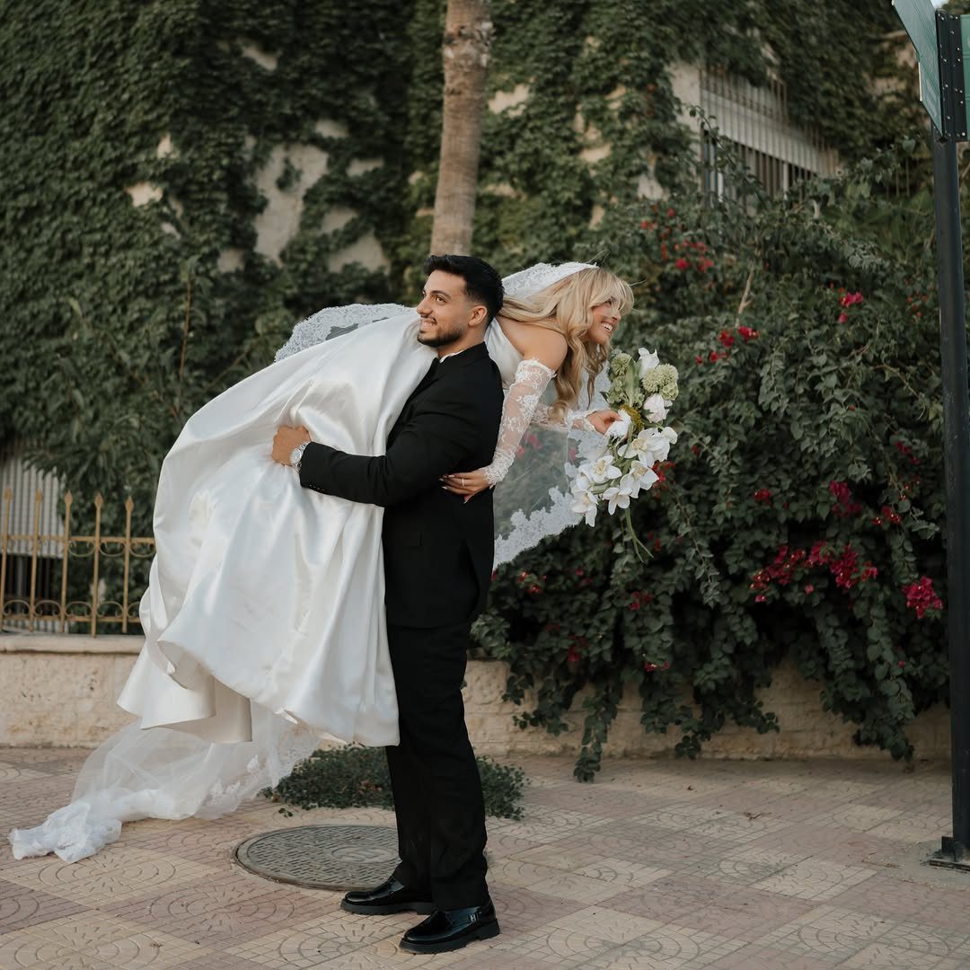 Groom carrying his bride in a white wedding dress outdoors near green ivy.