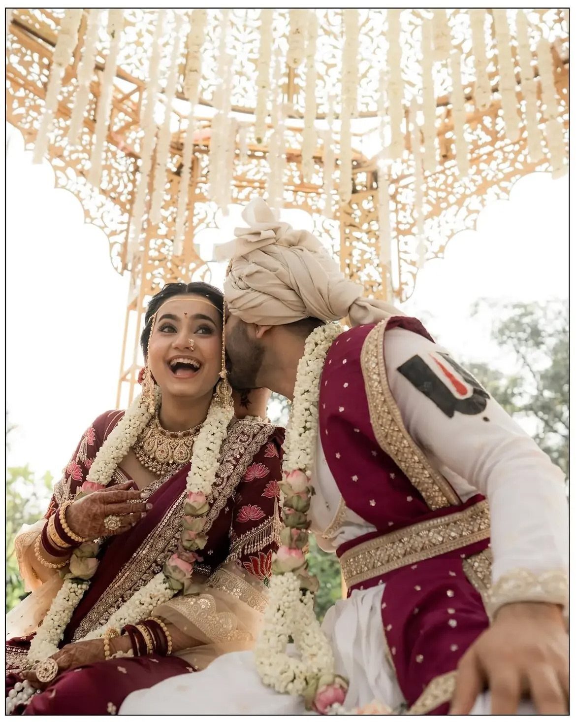 Laughing Indian bride and groom sharing a kiss under an ornate white canopy