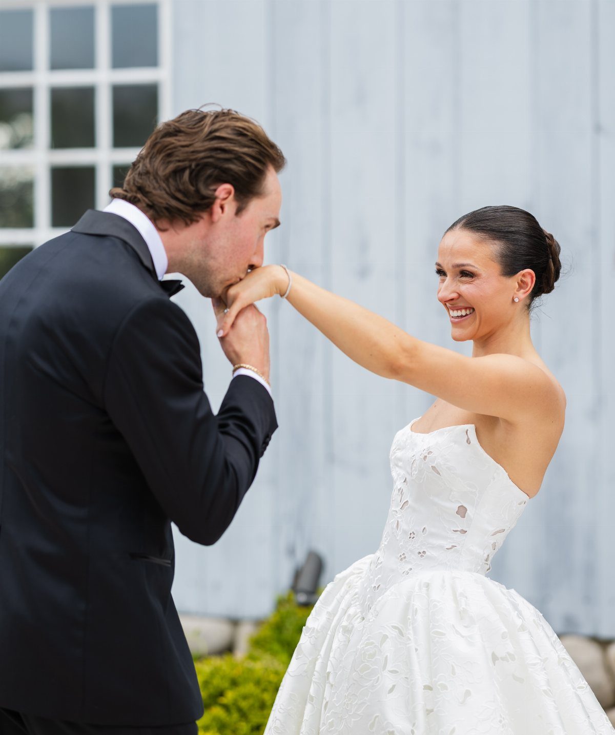 Groom in black tuxedo kissing the hand of a smiling bride in white gown