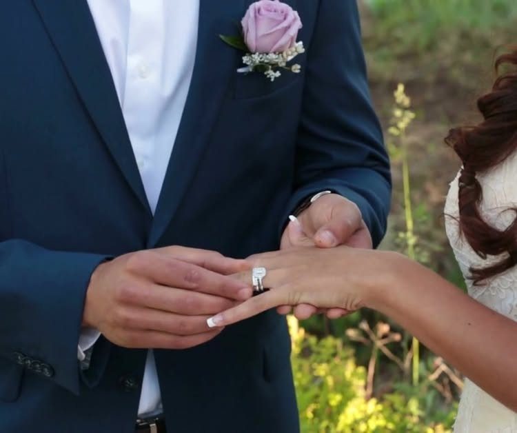 Groom in navy suit placing a wedding ring on the bride's finger outdoors.