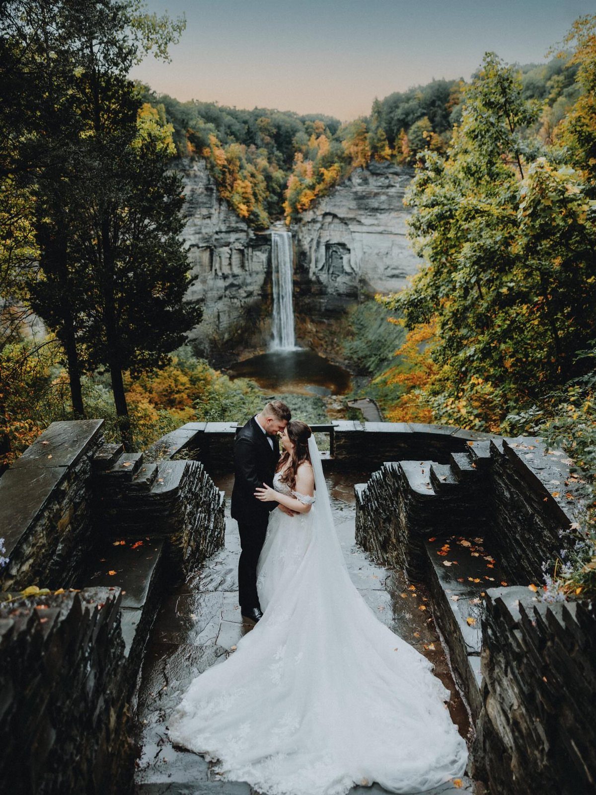 Bride and groom embracing on a stone terrace with a waterfall background.