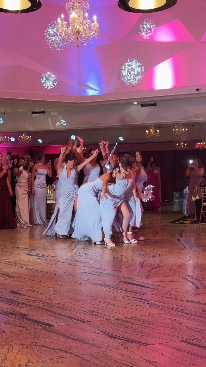 Bridesmaids in light blue dresses dancing on a ballroom floor under colorful lights.