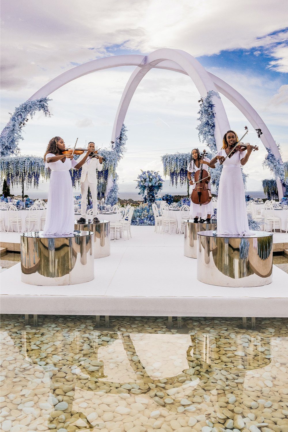 String quartet in white playing instruments on silver pedestals under white floral arches.