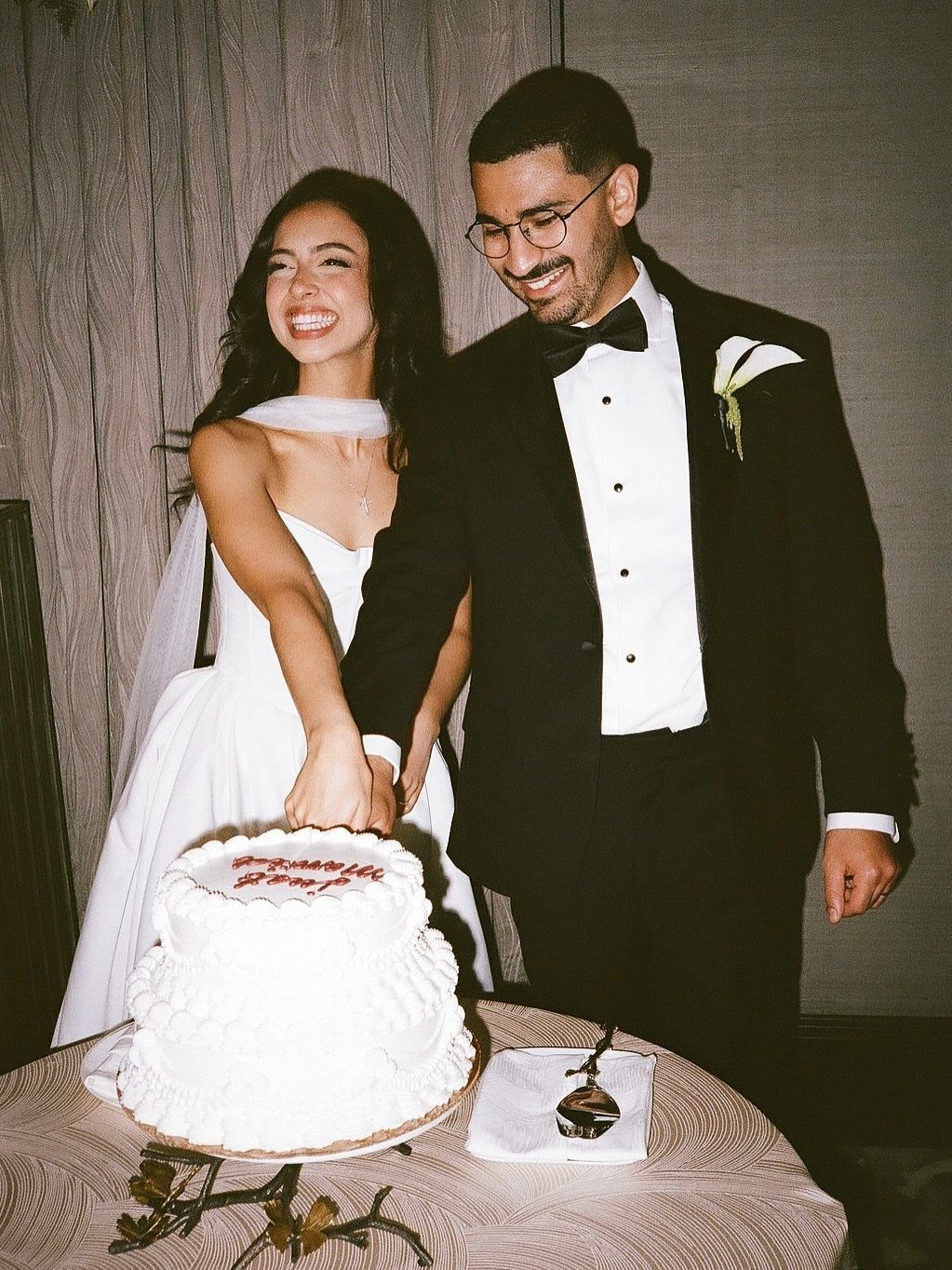 Bride and groom smiling while cutting a white tiered wedding cake together.