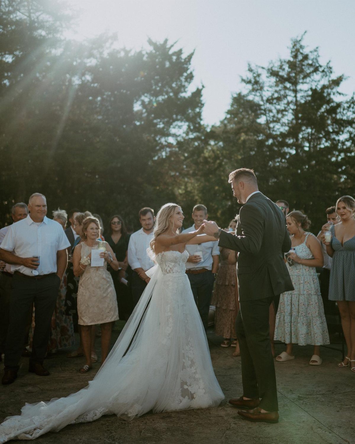 Bride and groom dancing outdoors surrounded by guests with bright sunlight filtering through trees