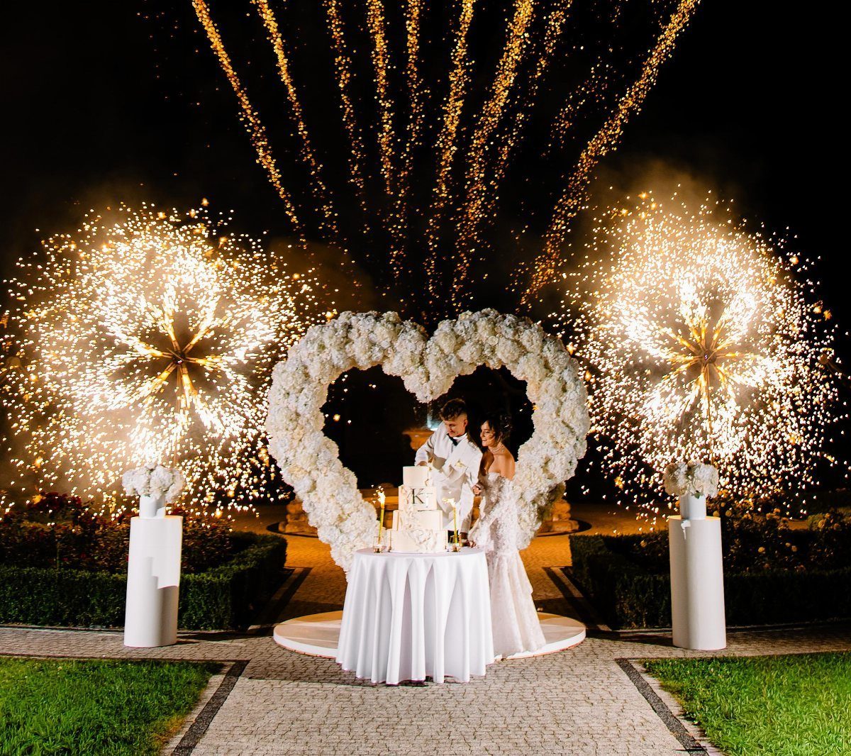 Bride and groom by a wedding cake under a floral heart with fireworks.