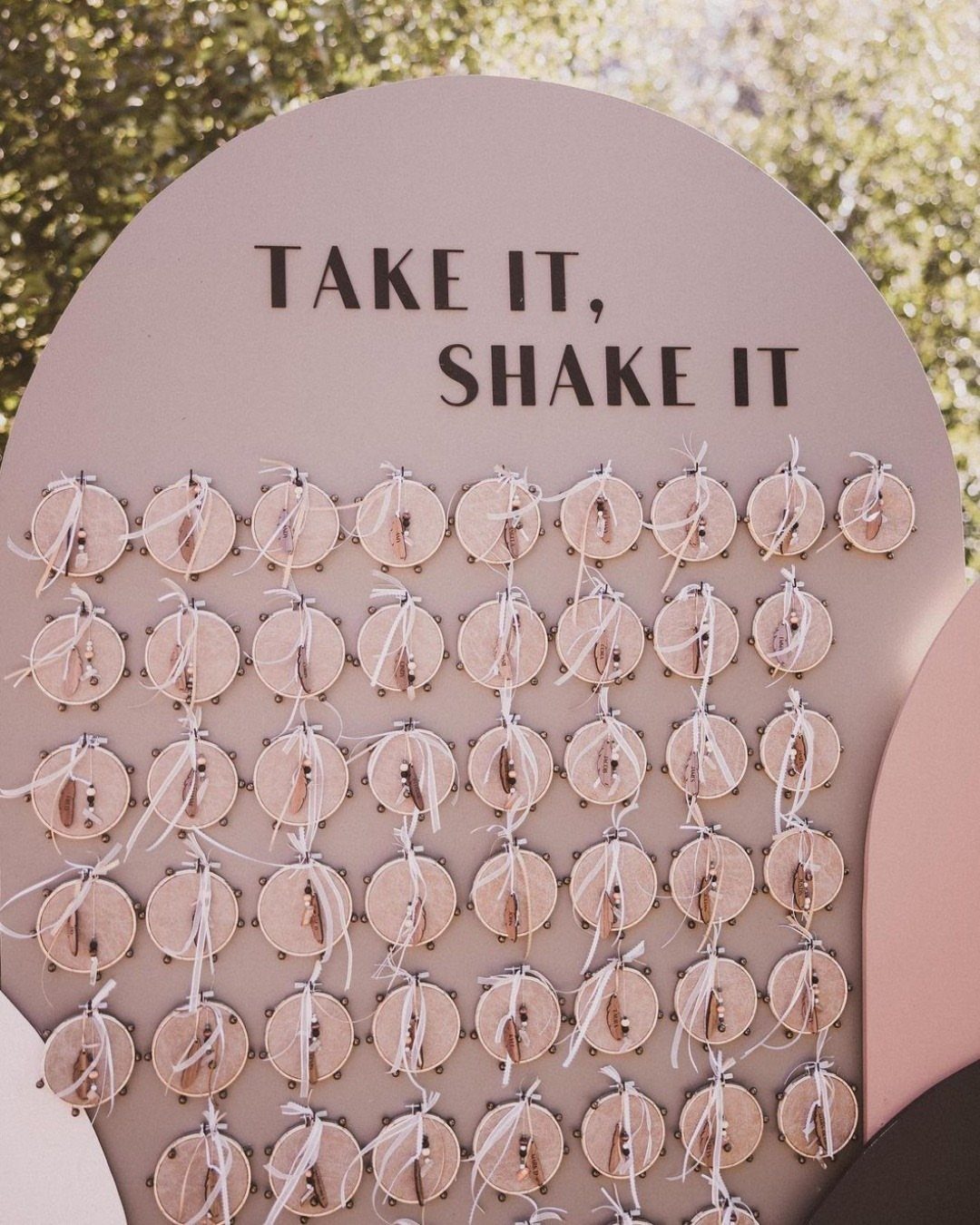 Pink arched board with bells and text 'TAKE IT, SHAKE IT' for wedding guests.