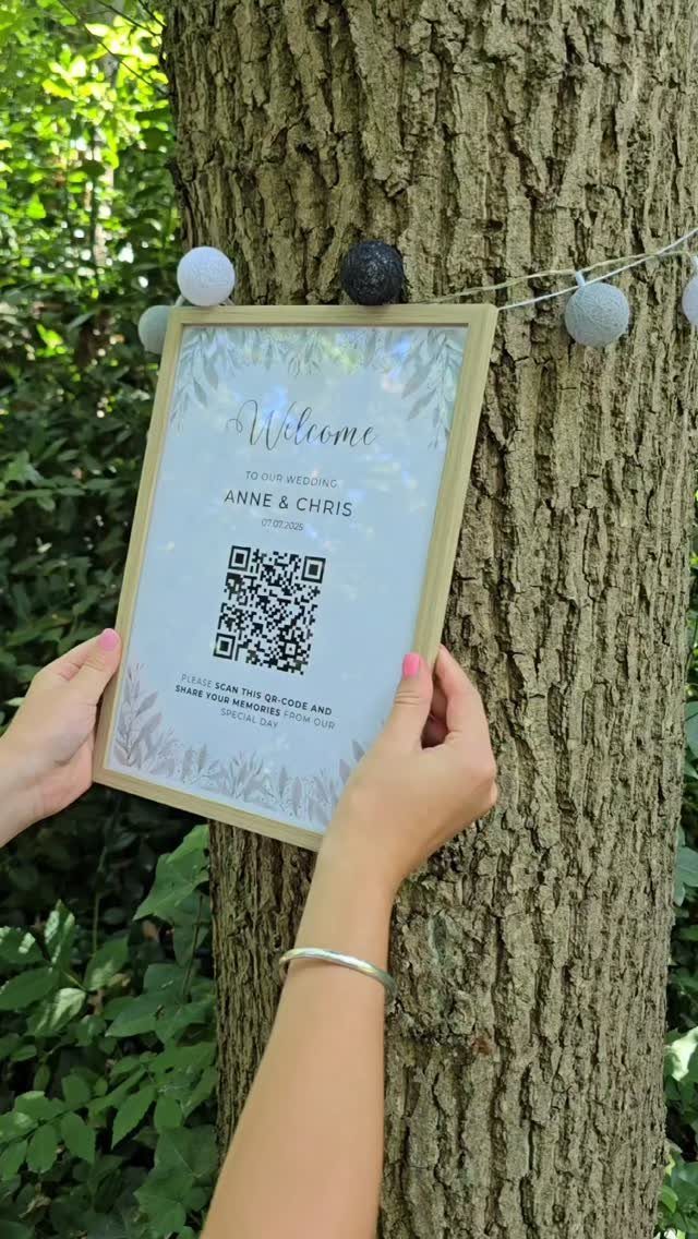 Gold-framed wedding welcome sign with QR code held against a textured tree trunk.