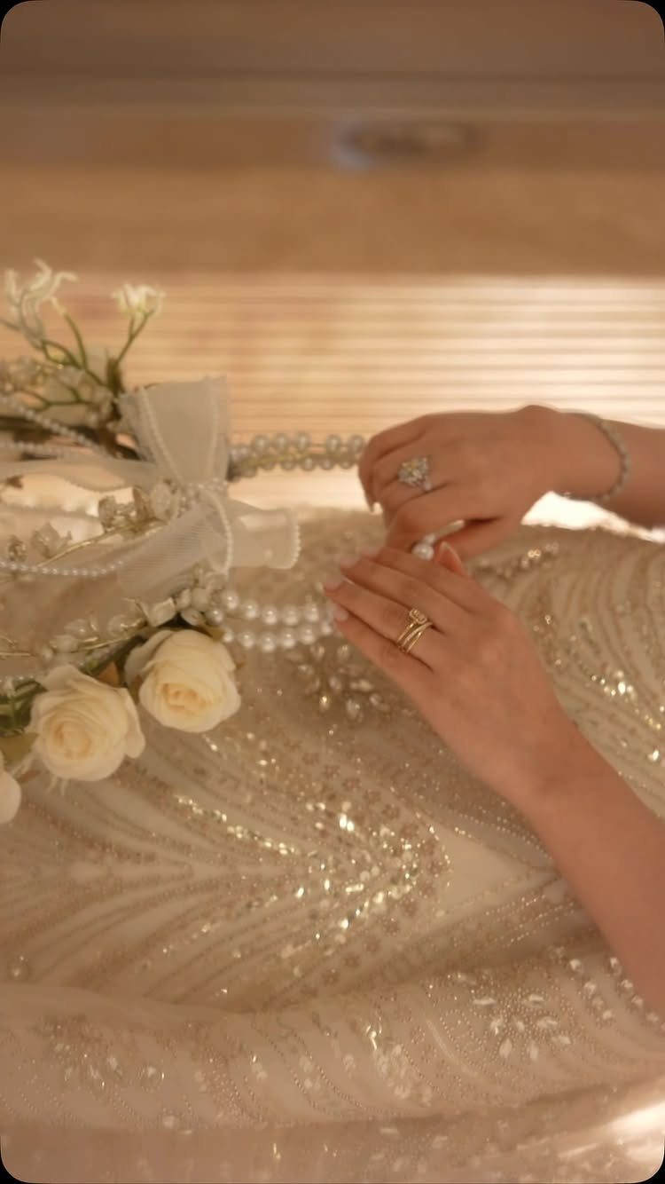 Hands with gold rings resting on a beaded white wedding dress with roses.